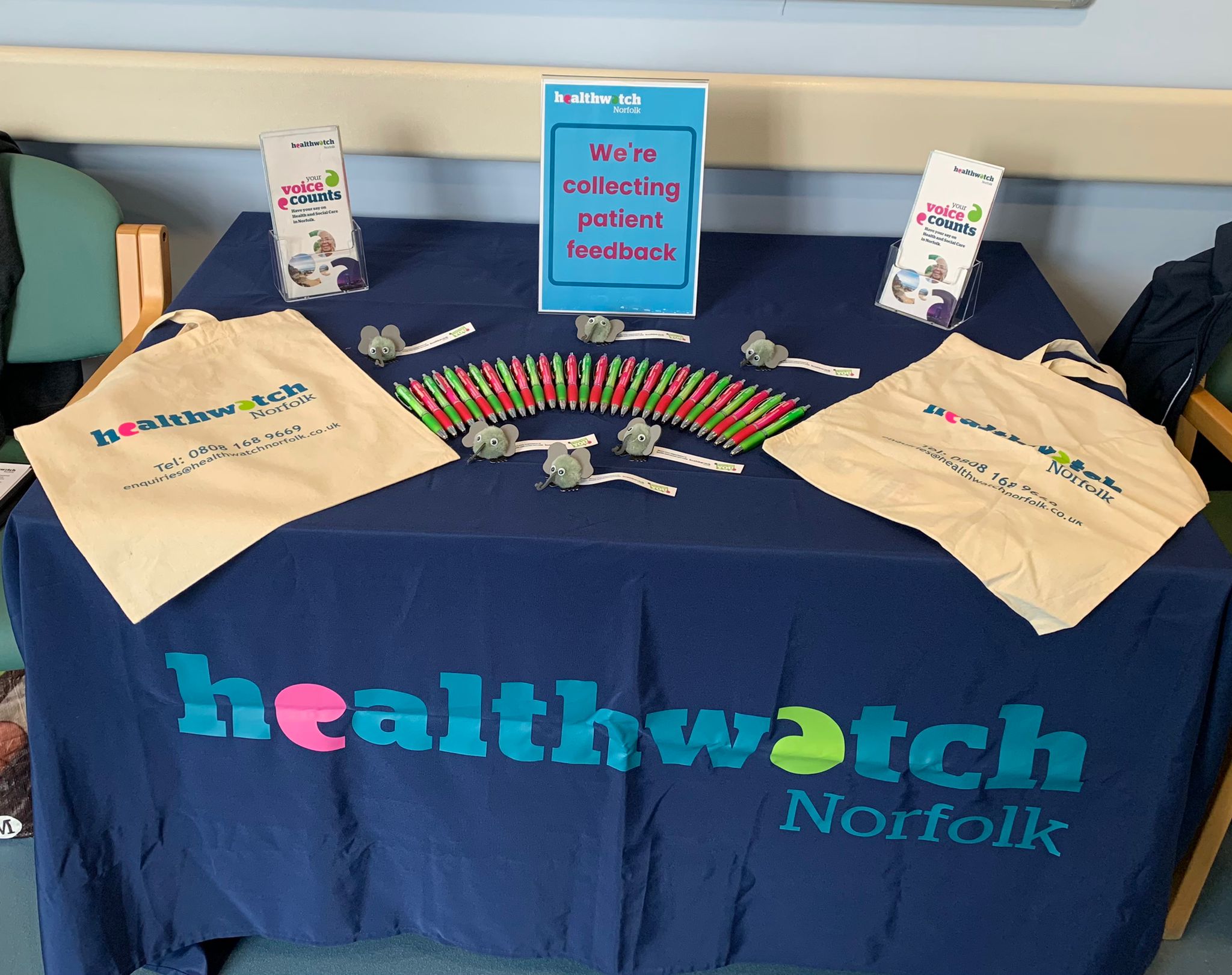 image shows a table with dark blue tablecloth and the words healthwatch norfolk printed on. On the table are two healthwatch bags, a fan of green and pink pens. There is a sign saying 'we are collecting patient feedback' and some leaflets