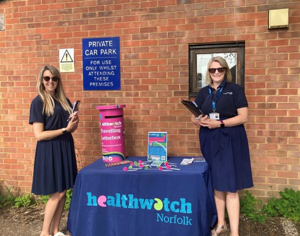 two people in healthwatch uniform standing next to a healthwatch branded table. On the table is some healthwatch pens, a sign saying we are collecting feedback and a small pink postbox